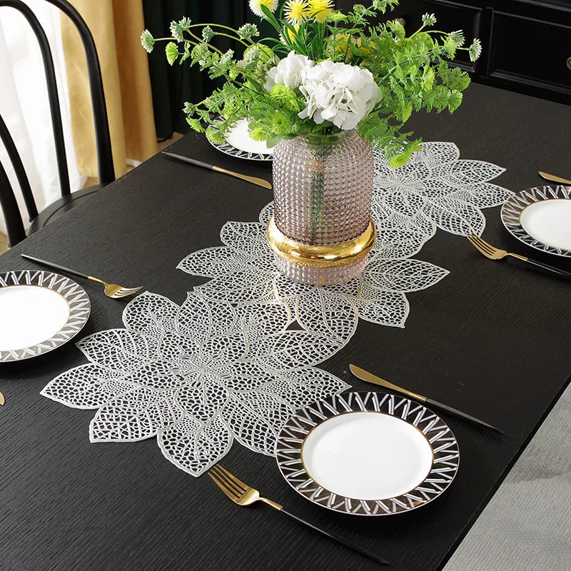 A black dining table set with gold cutlery, white plates, and decorative lace placemats and a floral centerpiece.
