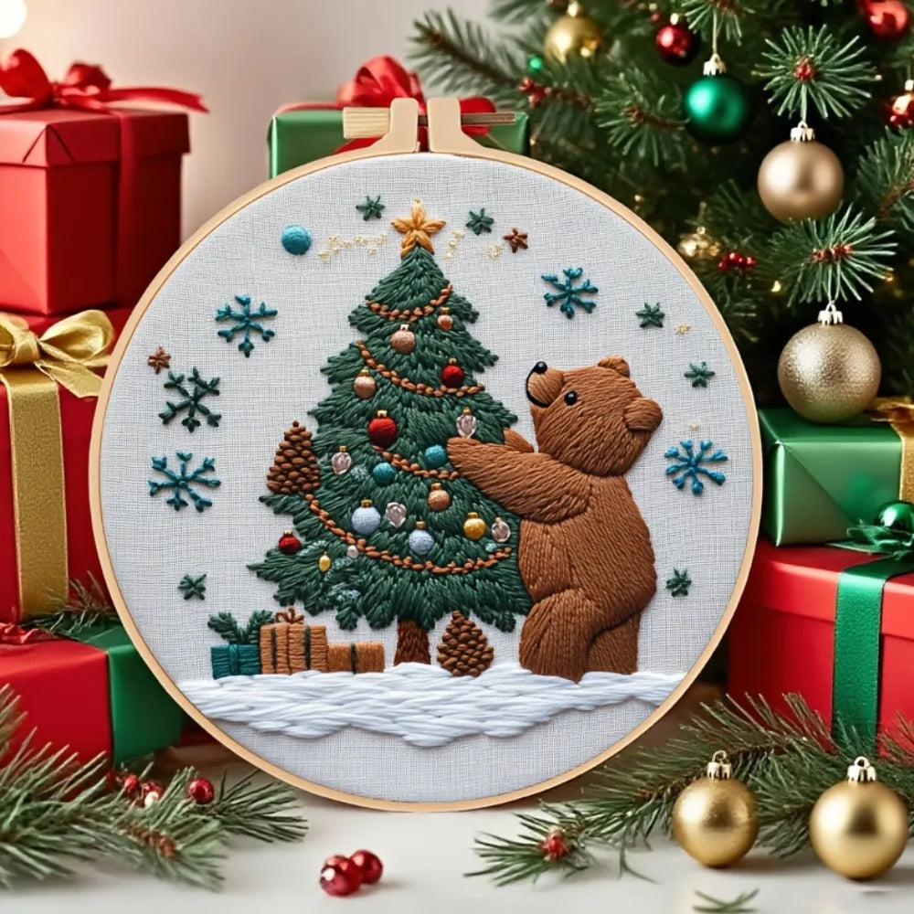 A hand embroidery kit featuring a stitched teddy bear and Christmas tree on a circular canvas surrounded by festive greenery 