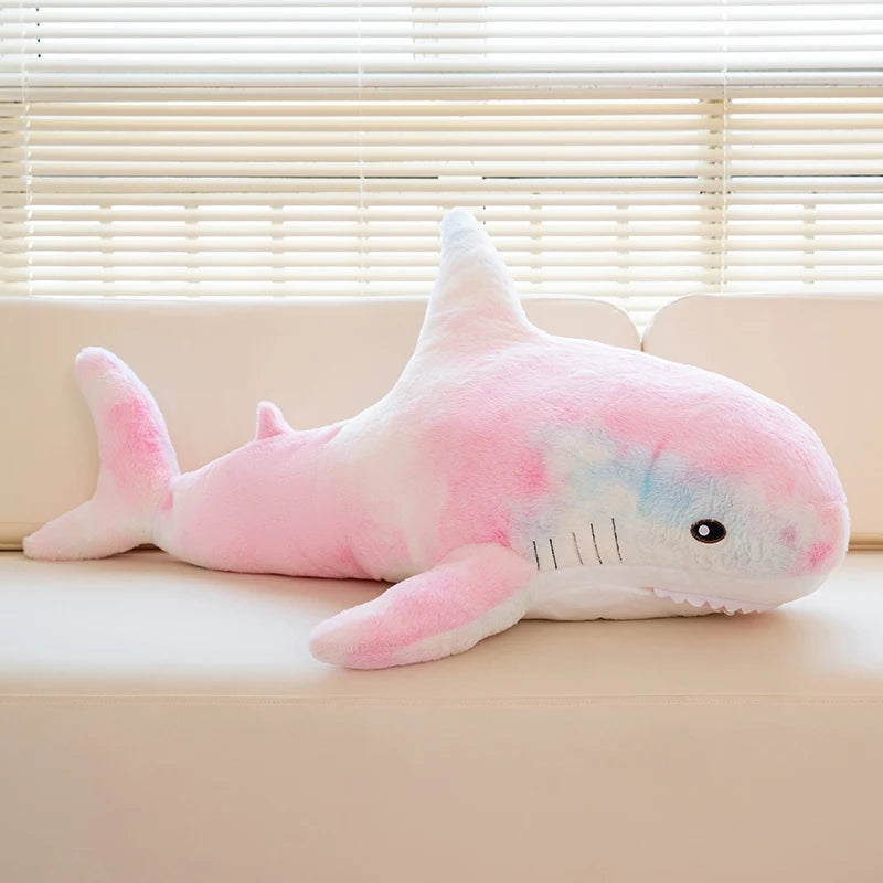 A pink and blue giant shark plush toy lying on a light-colored surface near a window.