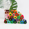A collection of 24 colored sewing threads in spools is displayed next to a green leaf and a brown glass bottle.