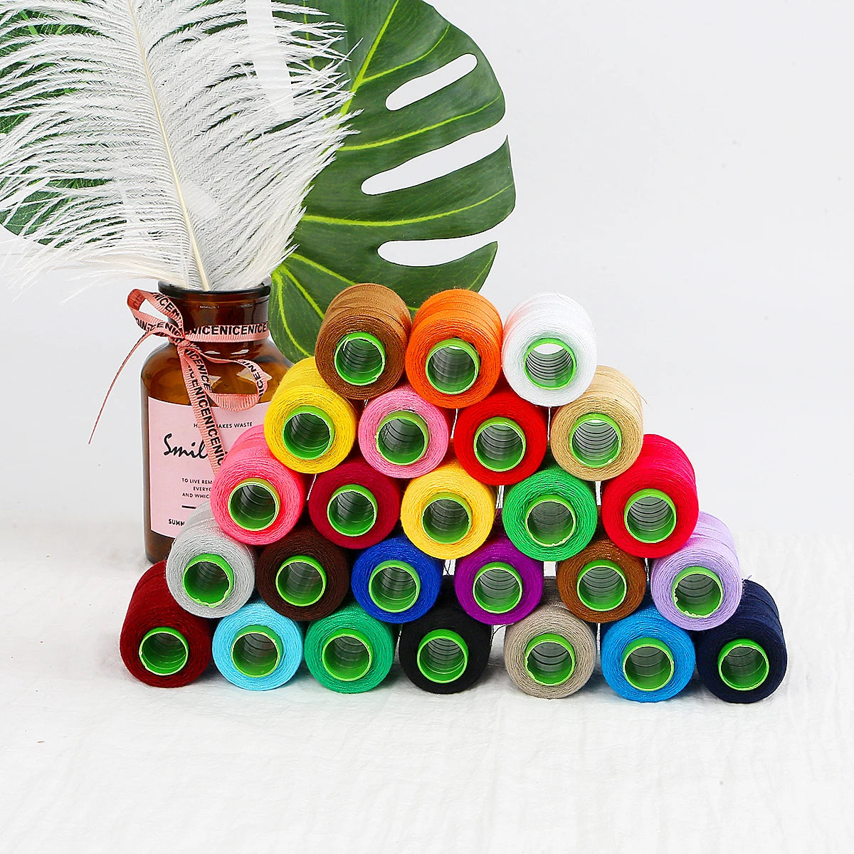 A collection of 24 colored sewing threads in spools is displayed next to a green leaf and a brown glass bottle.