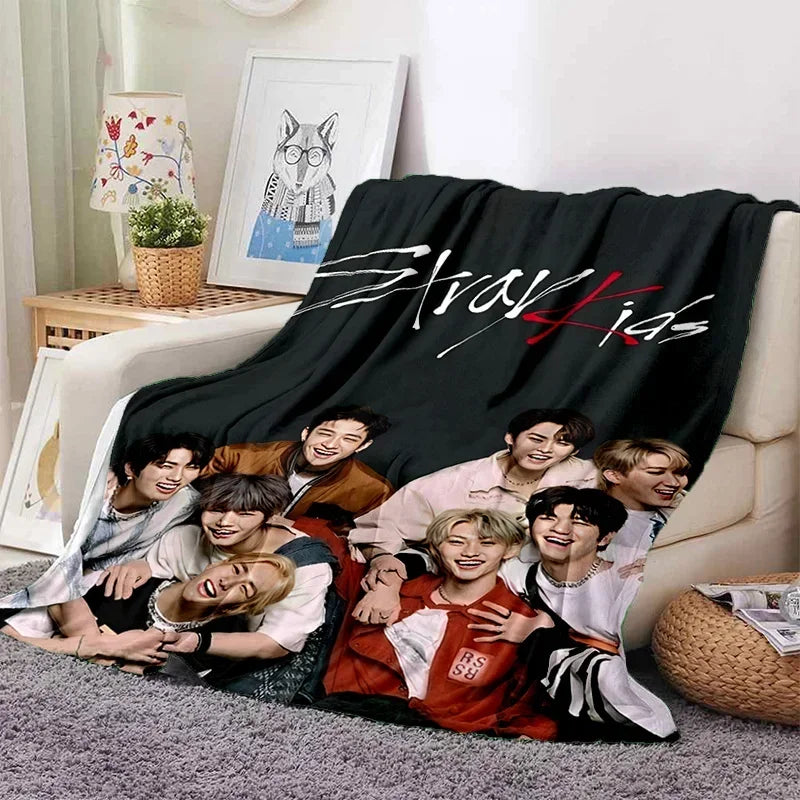 A black fleece throw blanket featuring the Stray Kids logo printed in red on a light beige couch.