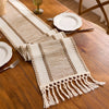 A beige macrame tassel table runner is laid out on a wooden table with silverware and a white plate.