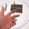 A hand holds a single black knitting needle with floral embroidery, next to a grid of similar needles.