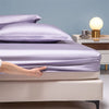 A hand gently touches a purple luxury bamboo bedding set on a bed with a light blue wall and bedside lamp.
