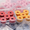 A collection of prewound sewing machine bobbin threads in various colors, including pink, red, and yellow, packaged in clear 
