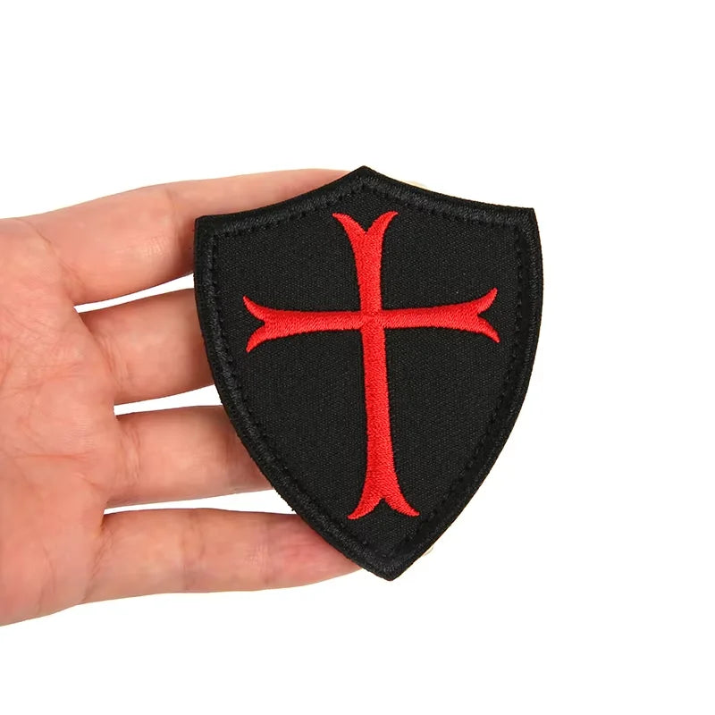A black shield-shaped patch embroidered with a red cross is held in a hand.