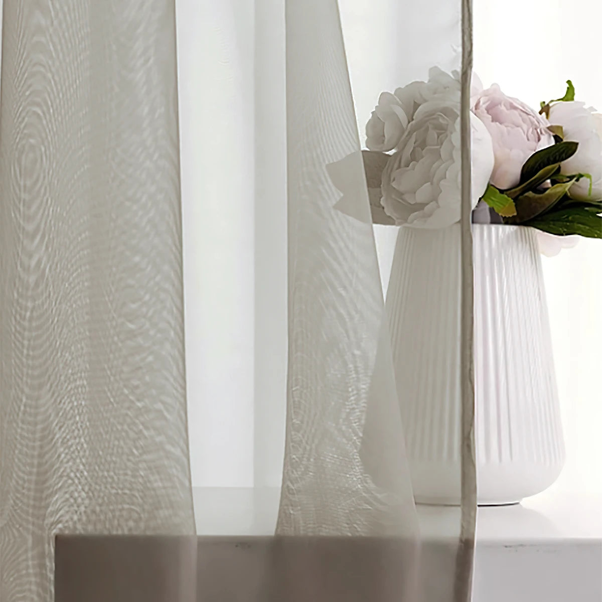 A close-up of a sheer white curtain with a rod pocket, draped next to a vase of pink and white flowers.