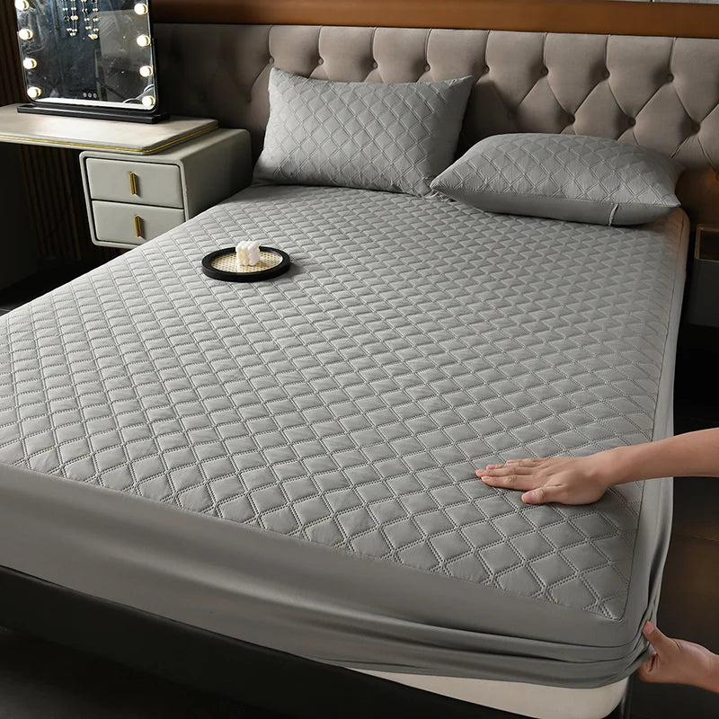 A gray quilted mattress protector is shown on a bed, with a decorative pillow and a candle on the surface.