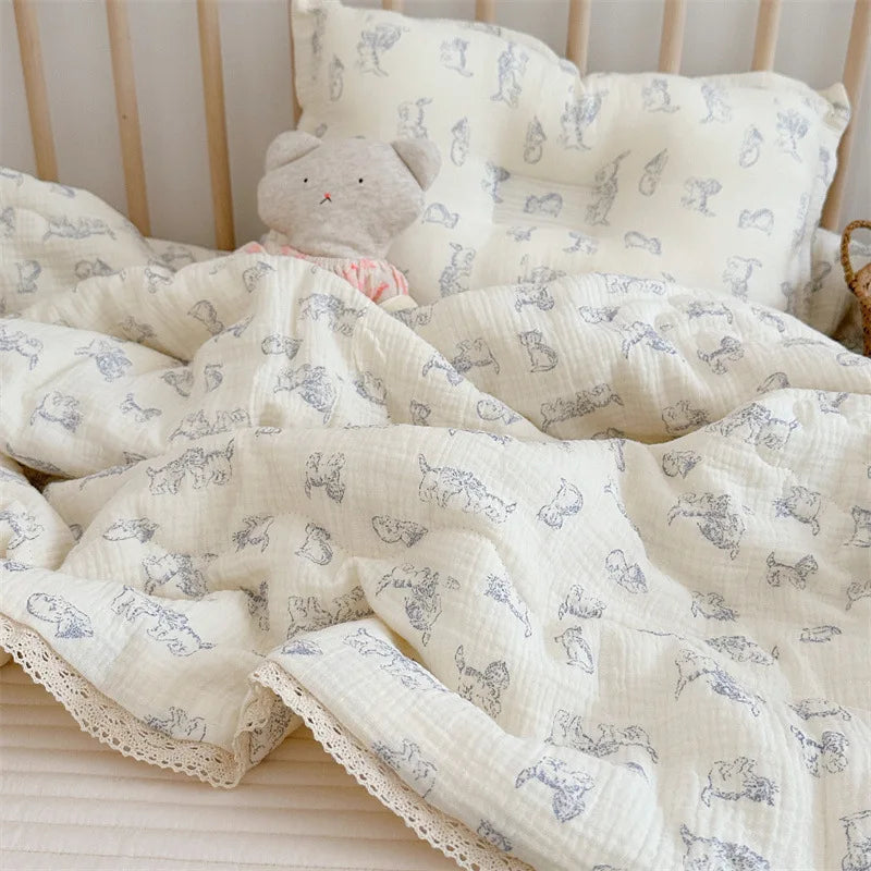 A white muslin blanket with a light blue floral print is draped over a bed and next to a stuffed animal pillow.