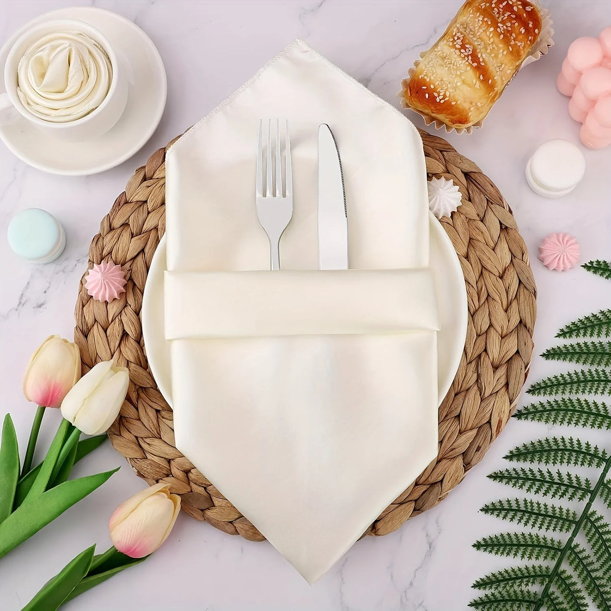 A set of silky satin dinner napkins is displayed on a wooden board with silverware and a green fern.