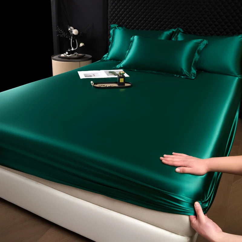 A close-up of a dark emerald green satin fitted sheet on a bed, with a hand touching the fabric.