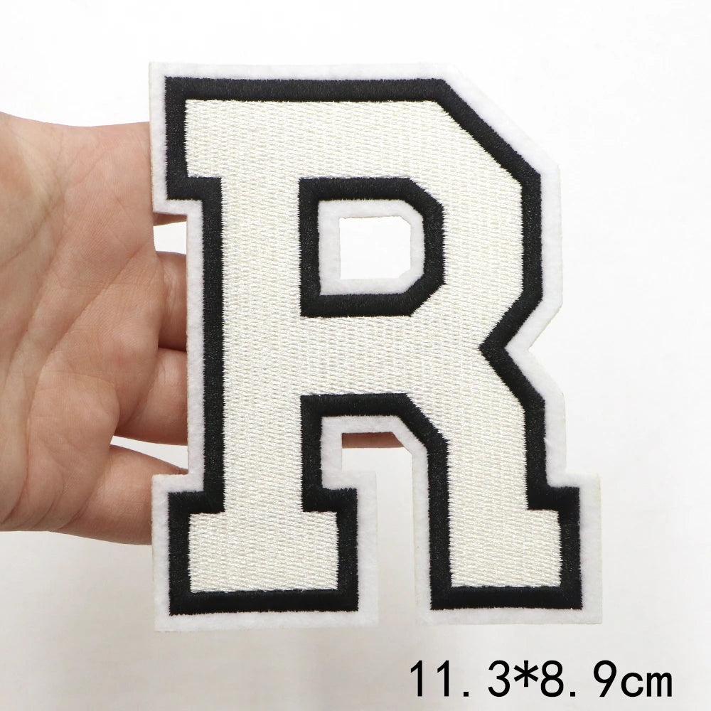 A hand holds a white letter R patch with black outline and stitching on a white background.