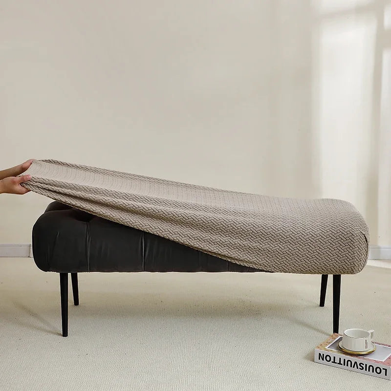 A beige elastic jacquard bench cover is draped over a dark upholstered bench in a bright room.