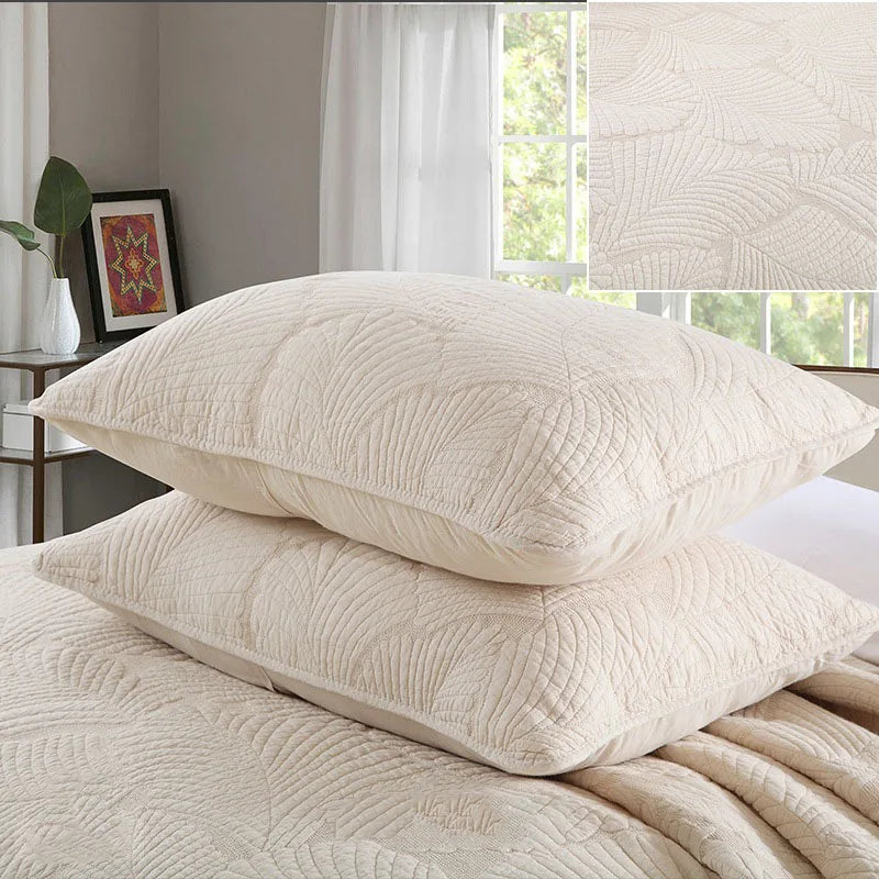 A set of cream embroidered cotton quilted pillow covers resting on a white bed with sheer curtains in the background.
