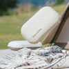 A white inflatable camping pillow is shown resting on a tan mat outdoors.