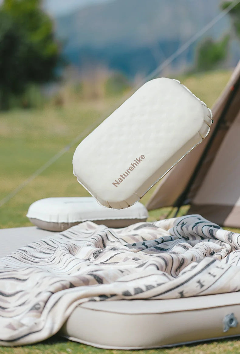 A white inflatable camping pillow is shown resting on a tan mat outdoors.