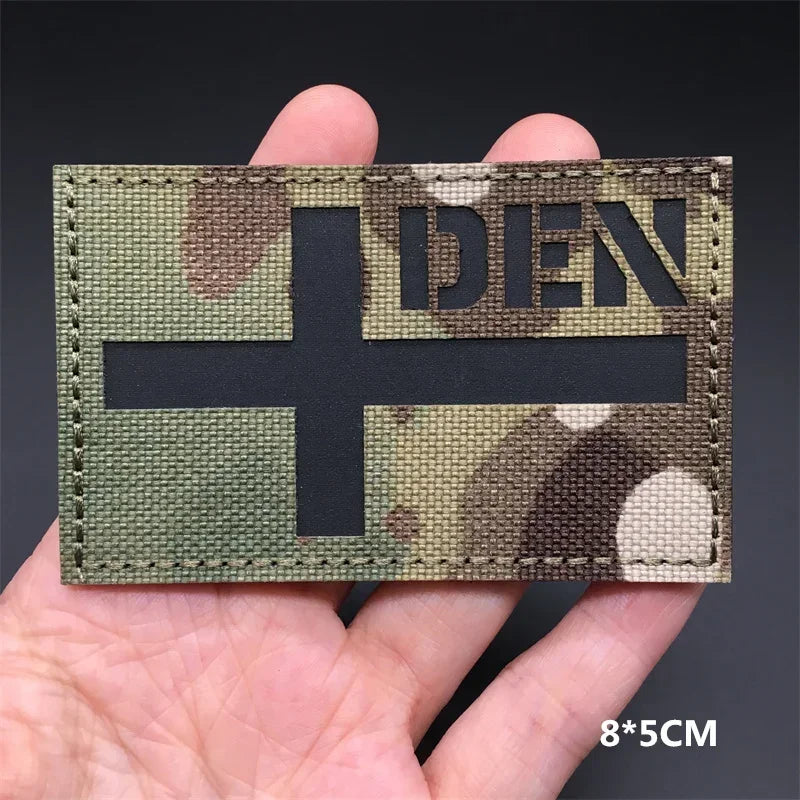 A rectangular tactical patch featuring a dark cross on a camouflage background held in a hand.