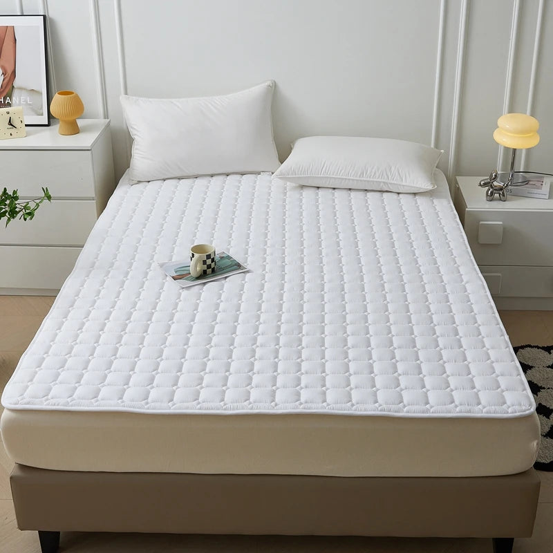 A white quilted mattress protector is displayed on a bed, next to bedside tables and pillows.