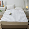 A white quilted mattress protector cover is displayed on a bed, next to bedside tables and pillows.