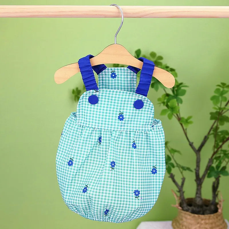 A light blue and green gingham dog dress with small blue flowers hanging from a wooden hanger against a green background.