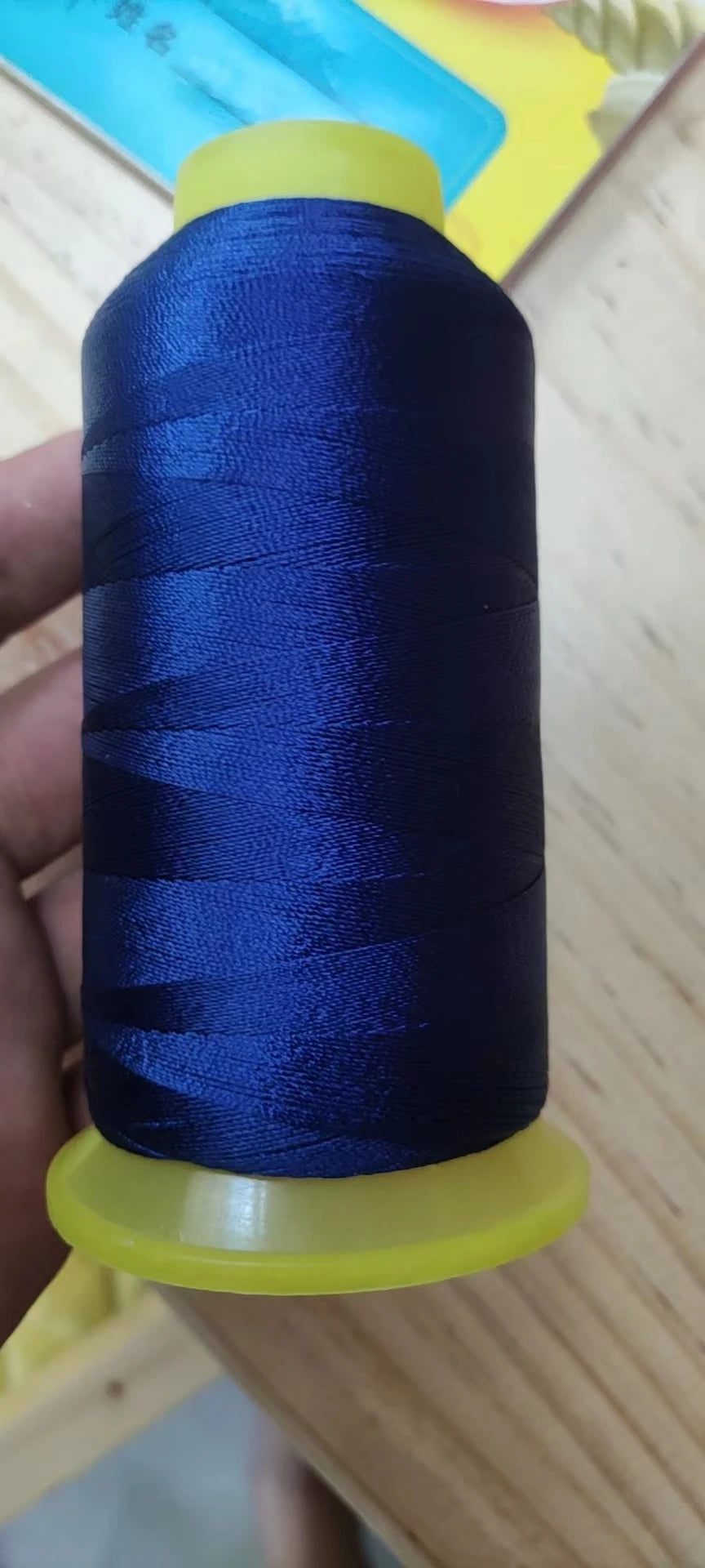 A spool of dark blue high tenacity polyester sewing thread with a yellow plastic core is held in a hand.