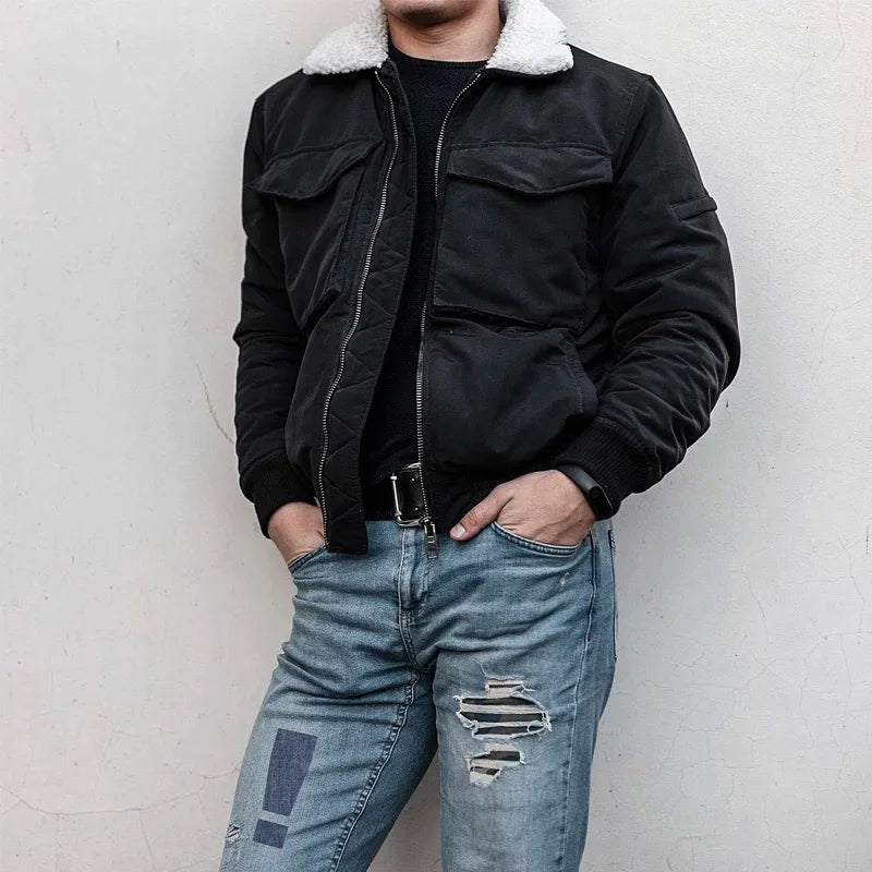 A person wearing a black jacket and jeans standing against a light wall.