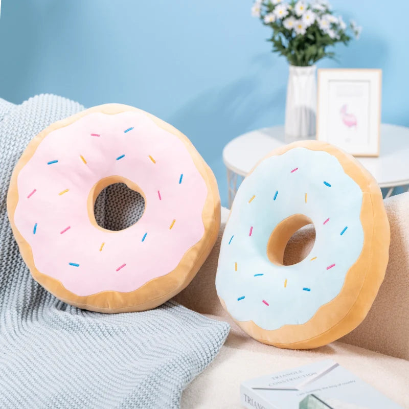 A pink and blue frosted doughnut plush pillow resting on a light gray knitted fabric.