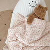 A fluffy brown teddy bear is resting on a pink floral print muslin blanket.