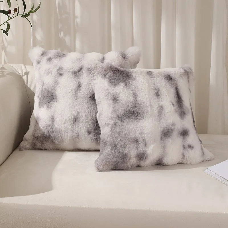 A pair of fluffy gray and white faux rabbit fur decorative throw pillows resting on a light-colored surface.