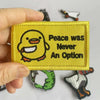 A hand holds a yellow embroidery patch featuring a smiling yellow chick and the text Peace was Never An Option.