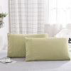 A set of two light olive green polyester pillow covers resting on a light gray surface near a window with a white grid curtai