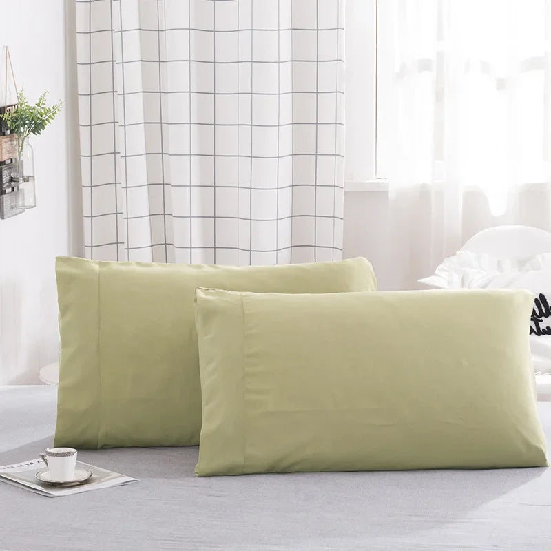 A set of two light olive green polyester pillow covers resting on a light gray surface near a window with a white grid curtai