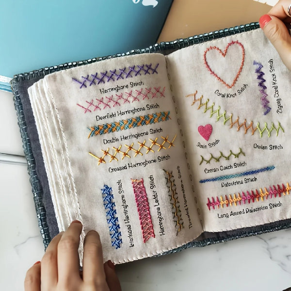 An open embroidery stitch book showing various hand embroidery stitches on a beige fabric.