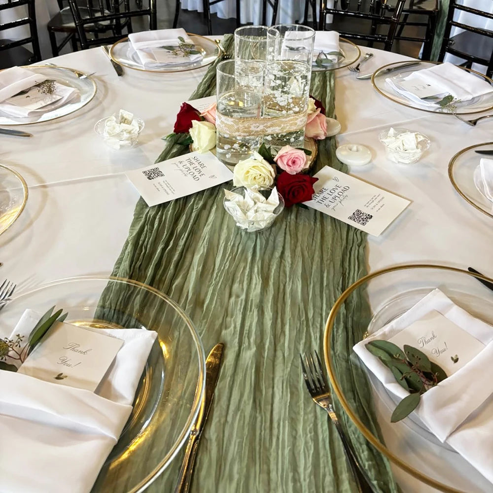 A long, sage green cheesecloth table runner is laid down on a white tablecloth for a rustic boho dining setting.