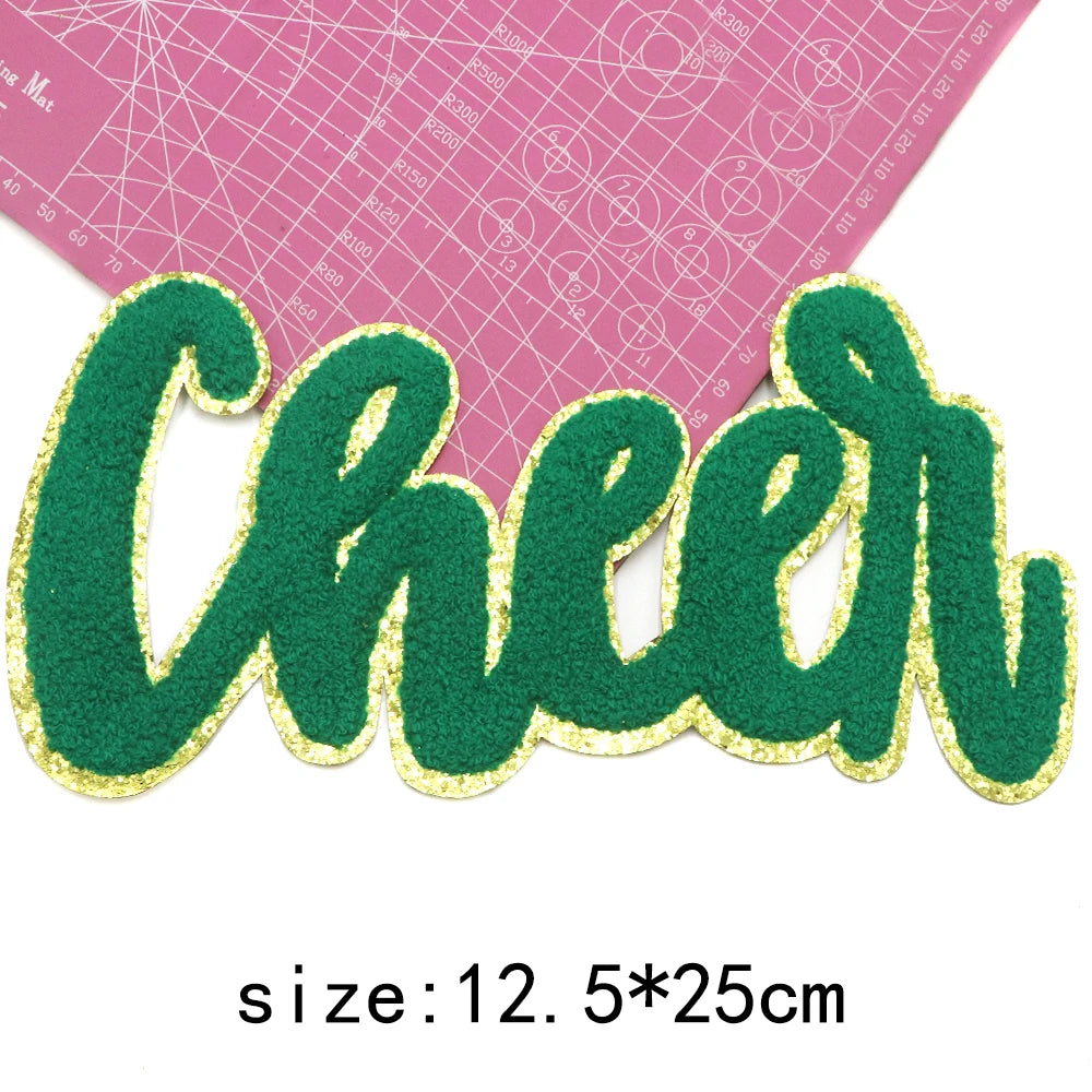 Green glitter script letter cheer iron-on patch on a pink grid background
