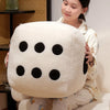 A woman is holding an oversized white dice plush throw pillow with black dots, sitting indoors.