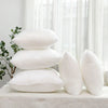 A stack of white cotton-filled decorative throw pillow inserts resting on a white surface near a window.