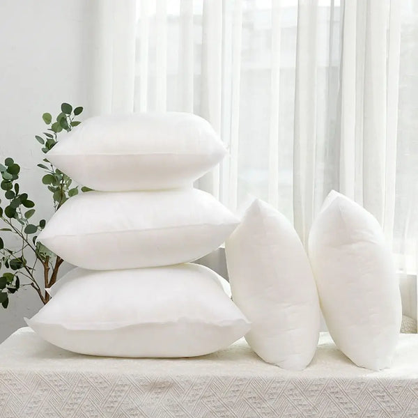 A stack of white cotton-filled decorative throw pillow inserts resting on a white surface near a window.