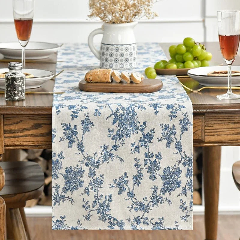 A blue floral patterned table runner on a wooden table set for dining with fruit and glassware.