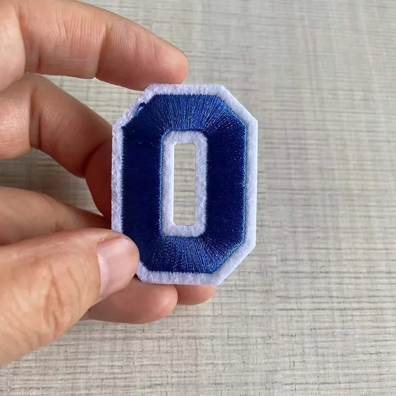A hand holds a dark blue felt number patch shaped like the numeral zero.