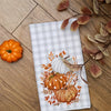 A white checkered kitchen towel featuring an orange and white pumpkin print on a wooden surface.