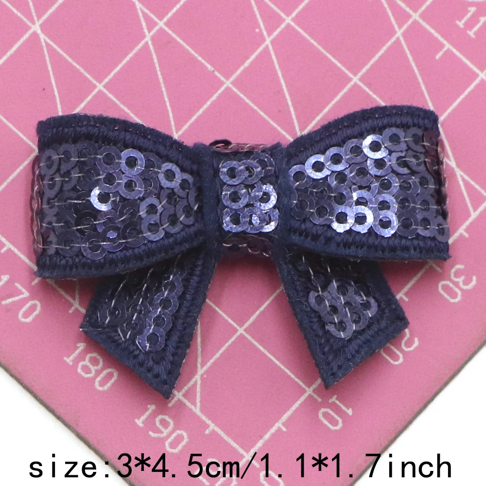 A dark purple sequin bow patch for DIY decoration shown against a pink grid background.