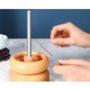 A hand is using a stainless steel beading needle to thread beads onto a wooden tool for crafting.
