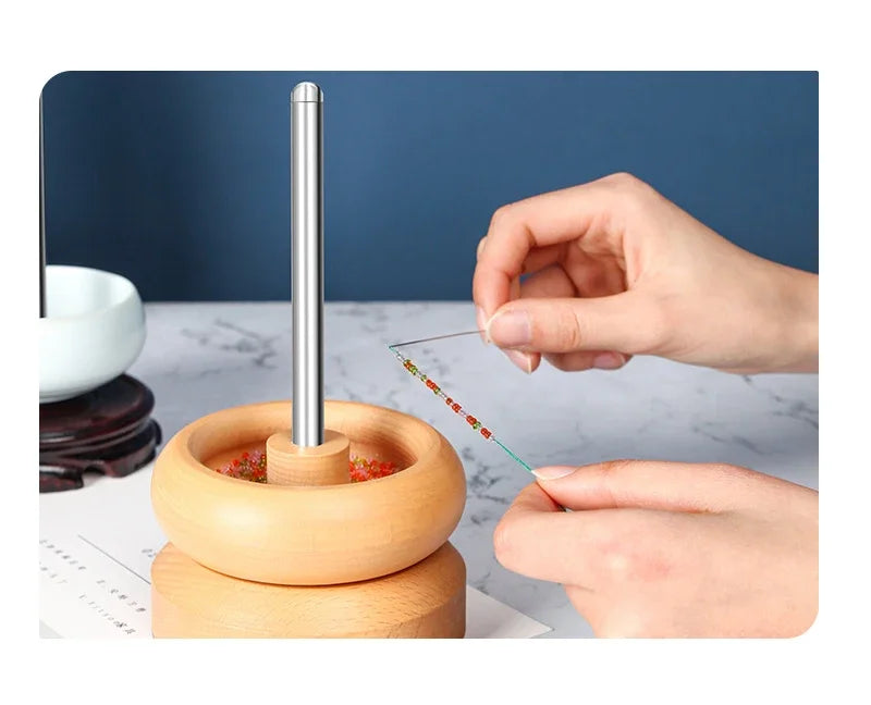 A hand is using a stainless steel beading needle to thread beads onto a wooden tool for crafting.