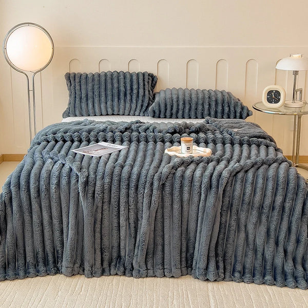A thick, gray faux fur blanket is draped over a bed, with matching pillows and a bedside lamp visible.