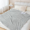 A light gray knitted throw blanket draped over a white bed with soft pillows and sheer curtains.