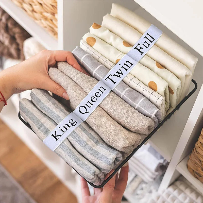 A hand holds a stack of four folded linen bed sheets tied with elastic straps, labeled King and Queen sizes.