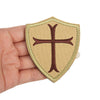 A hand holds an embroidered military cross shield patch with a tan background and brown cross design.