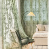 A sheer floral lace curtain panel in sage green hangs next to a window with slatted blinds and a modern lamp.